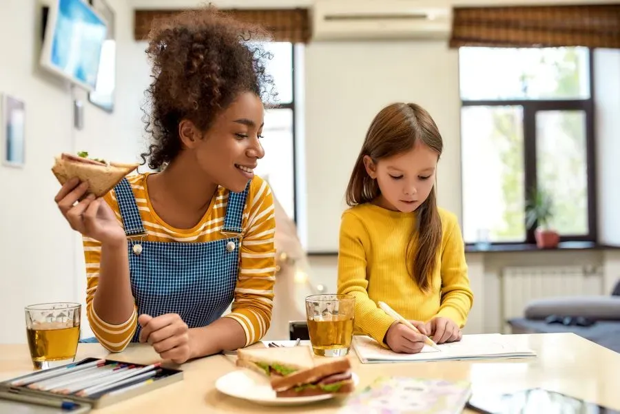 Woman in overall with yellow shirt holding a sandwich watching a little girl in a yellow shirt write on a piece of paper, doing homework. part of what is happening during the hot Southwest Florida weekend